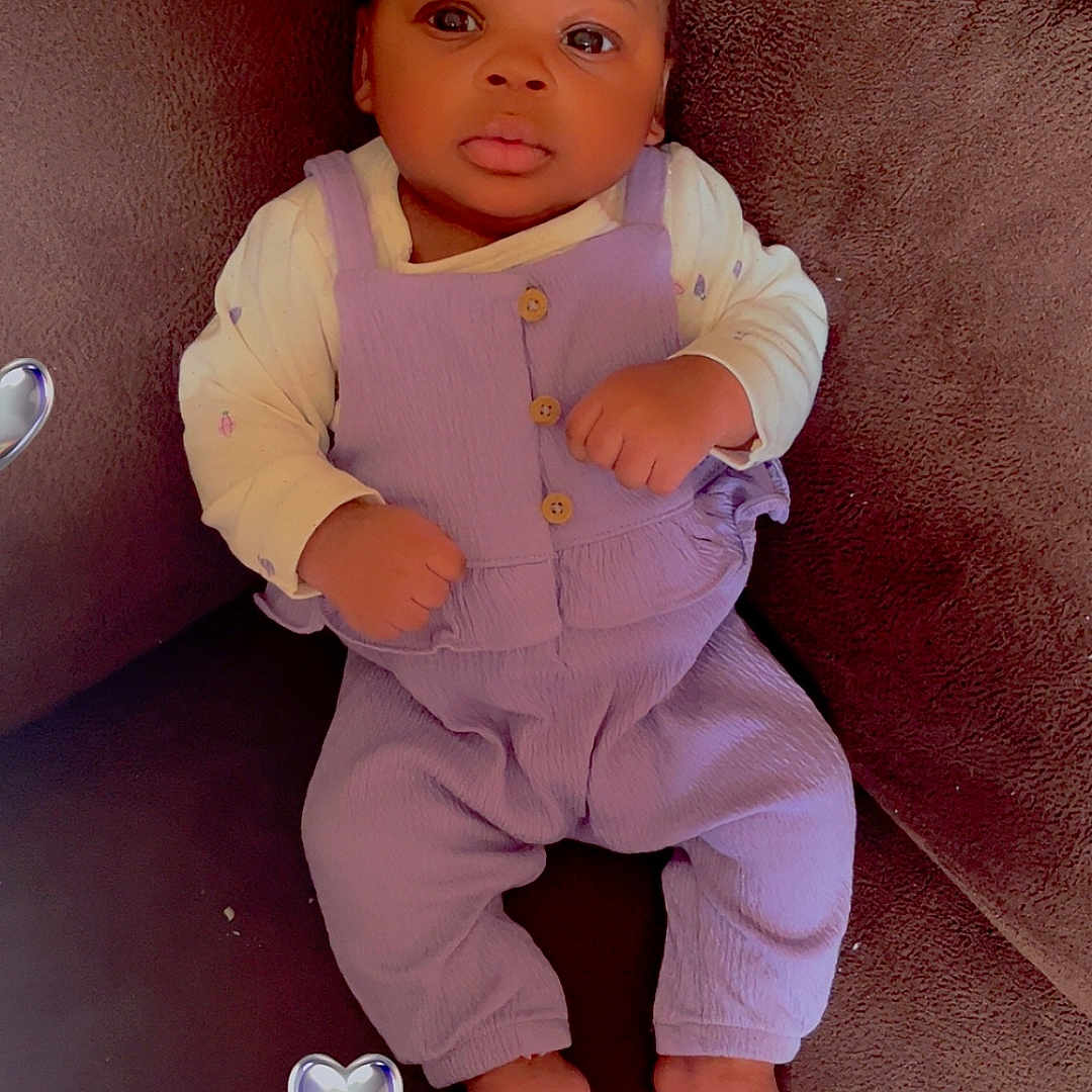 Kanyla is registered to the contest to win money with this photo: baby, infant, purple_overalls, cream_shirt, brown_couch, face, barefoot, heart_sticker, indoor, child, cute, portrait, looking_at_camera, relaxed, seated, hands_clenched, dark_skin, short_hair, soft_lighting, cozy