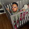 Ariyah joined the competition — help win amazing prizes! toddler, child, crib, glasses, headband, blanket, teddy_bear, wooden_floor, indoor, baby, furniture, person, standing, curious, pink_glasses, gray_crib, smile, baby_clothes, handrail, room