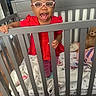 Ariyah joined the competition — help win amazing prizes! toddler, child, glasses, crib, blanket, toy, teddy_bear, red_clothing, pink_glasses, wooden_floor, indoor, smiling, happy, baby, nursery, furniture, person, standing, playful, cozy