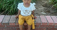 Ariyah is registered to the contest to win money with this photo: toddler, child, outdoor, greenery, plants, brick, shorts, white_shirt, sitting, curious, face, person, nature, casual_clothing, daylight, footwear, black_shoes, sidewalk, expression, young_child