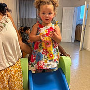 Aphora is registered to the contest to win money with this photo: toddler, child, curly_hair, floral_dress, slide, indoor, playroom, adult, supporting, floor, door, curtains, window, person, furniture, casual, expression, young_child, colorful_clothing, play