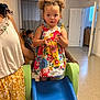 toddler, child, curly_hair, floral_dress, slide, indoor, playroom, adult, supporting, floor, door, curtains, window, person, furniture, casual, expression, young_child, colorful_clothing, play