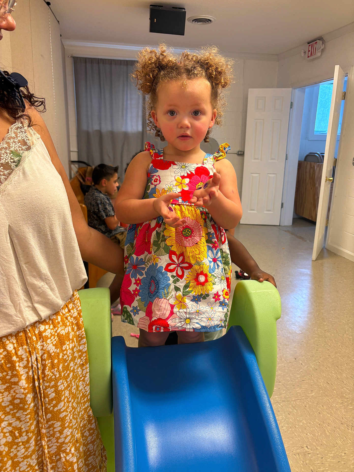 Aphora is registered to the contest to win money with this photo: toddler, child, curly_hair, floral_dress, slide, indoor, playroom, adult, supporting, floor, door, curtains, window, person, furniture, casual, expression, young_child, colorful_clothing, play