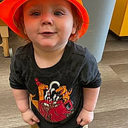 Larry is registered to the contest to win money with this photo: toddler, child, orange_hat, curious_expression, indoor, wooden_floor, trash_can, yellow_cabinet, casual_clothing, sneakers, black_shirt, pants, standing, looking_up, person, cute, young_child, funny_hat, playful, portrait
