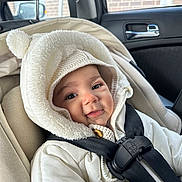 Eliana is registered to the contest to win money with this photo: baby, child, car_seat, white_jacket, hood, smile, straps, seatbelt, car_interior, window, face, cute, warm_clothing, infant, safety, headwear, button, black_straps, person, cozy