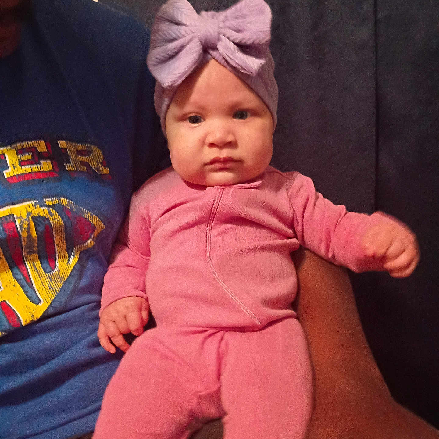 Anaya joined the competition — help win amazing prizes! baby, child, infant, portrait, sitting, being_held, face, headband, bow, pink_clothing, jumpsuit, serious_expression, indoor, close_up, person, adult_arm, blue_shirt, dark_background, fashion, photography