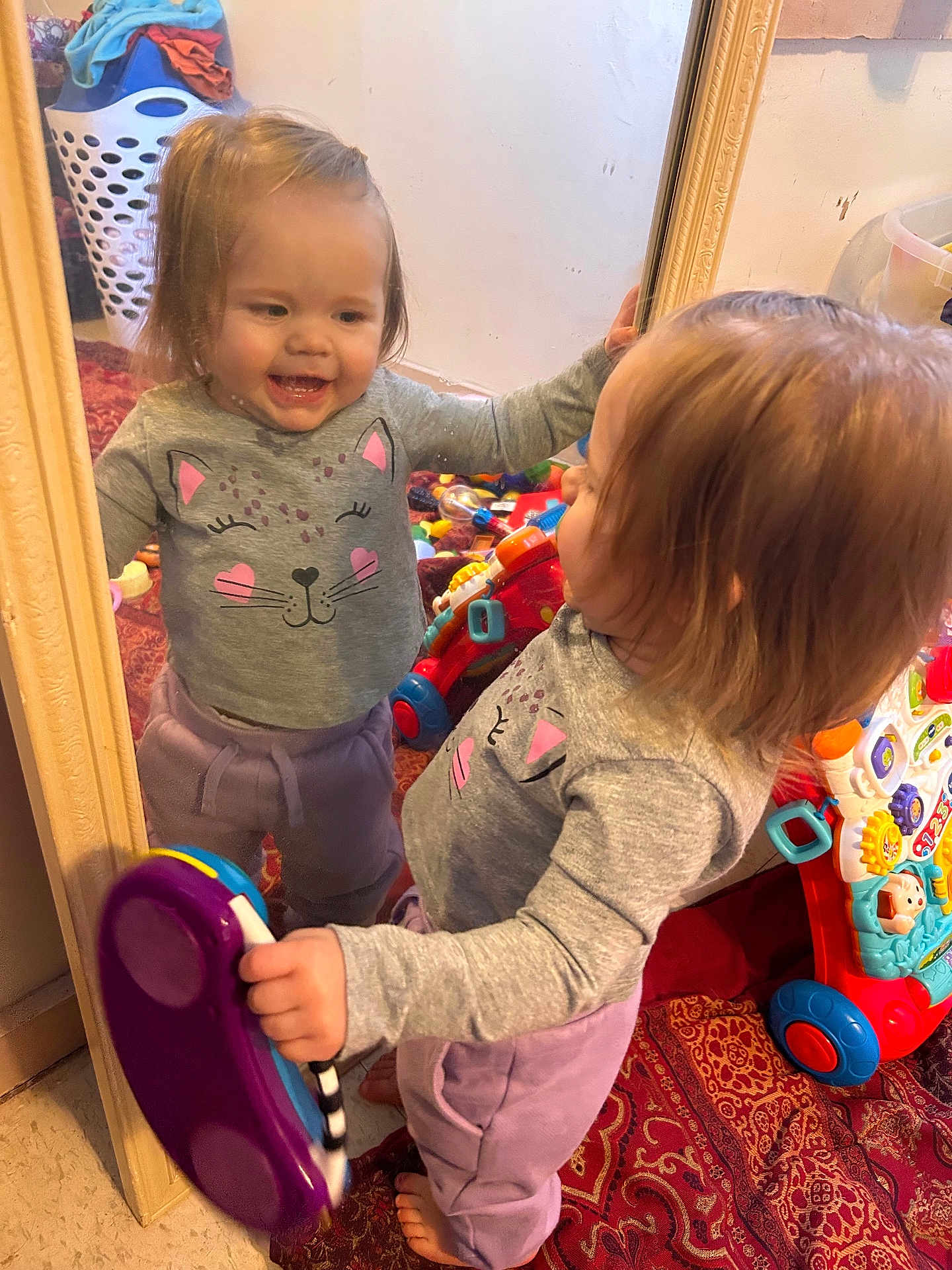 Lorelai Rain joined the competition — help win amazing prizes! toddler, child, mirror, reflection, toy, phone, gray_sweater, cat_face, purple_pants, barefoot, carpet, indoor, laundry_basket, smiling, happy, playing, furniture, room, person, home