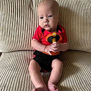 Abel is registered to the contest to win money with this photo: baby, infant, child, couch, furniture, clothing, onesie, red, black, skin, feet, hands, face, portrait, indoors, texture, sitting, cute, expression, young