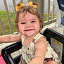 Natalie is registered to the contest to win money with this photo: arm, baby, child, curly_hair, cute, daylight, face, foot, happy, high_chair, outdoor, person, porch, railing, seat, smiling, summer_dress, table, toddler, yellow_bows