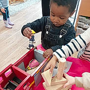 Kayden joined the competition — help win amazing prizes! child, toddler, toy, fire_truck, wooden_blocks, table, indoor, play, hands, denim_overalls, flooring, shelf, toy_figure, pink_tablecloth, person, playroom, casual_clothing, building_blocks, wooden_toys, focused