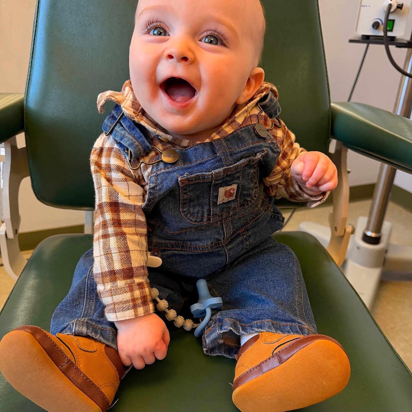 Seamus is registered to the contest to win money with this photo: baby, child, smile, happy, denim_overalls, plaid_shirt, brown_shoes, green_chair, indoor, person, cute, infant, sitting, clothing, footwear, expression, young_child, portrait, room, joyful