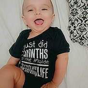 Jaxson is registered to the contest to win money with this photo: baby, child, smiling, mohawk, onesie, black_clothing, white_text, lying_down, bed, blanket, happy, infant, cute, portrait, indoor, skin, hand, foot, face, joyful
