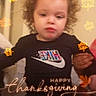 child, curly_hair, black_shirt, nike_logo, indoor, thanksgiving, autumn, leaf_graphics, holiday, festive, person, cute, young_child, blurry, celebration, decorations, hand, holding, casual_clothing, portrait