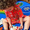 child, toddler, curly_hair, red_pajamas, sneakers, picture_book, play_mat, animal_pattern, indoor, wooden_floor, colorful, toy, reading, sitting, floor, casual, cute, young_child, playtime, comfortable
