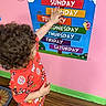 child, curly_hair, red_pajamas, poster, days_of_the_week, pink_wall, wooden_floor, green_baseboard, indoor, pointing, educational, colorful, footwear, black_shoes, patterned_clothing, person, standing, learning, youth, home