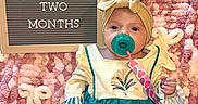 Anslee is registered to the contest to win money with this photo: baby, infant, pacifier, headband, blanket, sign, text, two_months, clothing, cute, pink, white, pattern, sitting, portrait, child, young, indoors, soft, comfort