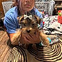 person, dog, indoor, blue_shirt, brown_hair, small_dog, bow, hands, furniture, water_bottle, blanket, wooden_floor, bracelet, table, curtains, pet, puppy, face, holding, home