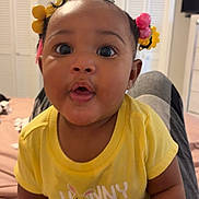 Jalaiah joined the competition — help win amazing prizes! baby, child, yellow_shirt, curly_hair, hair_bows, indoors, bedroom, person, cute, curious, wide_eyes, sitting, legs, toddler, clothing, face, expression, home, human, infant