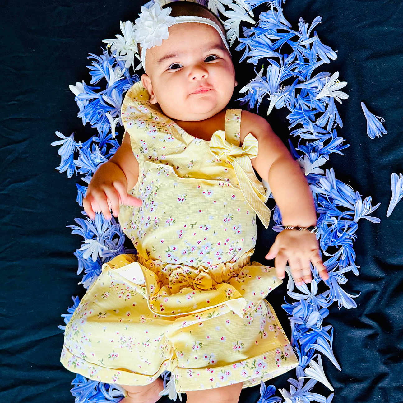 Erika is registered to the contest to win money with this photo: adorable, baby, barefoot, black_background, blue_flowers, child, curious_expression, cute, floral_pattern, flower_wreath, headband, infant, lying_down, outdoor_style, portrait, small_baby, smiling, soft_lighting, white_flowers, yellow_outfit
