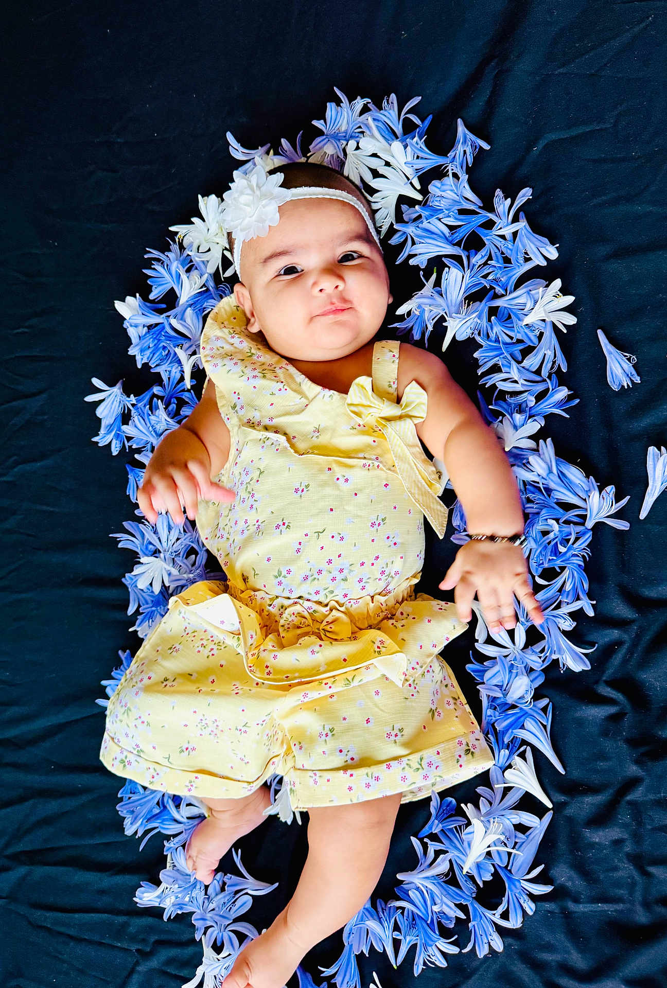 Erika is registered to the contest to win money with this photo: baby, child, flower_wreath, yellow_outfit, headband, blue_flowers, white_flowers, lying_down, curious_expression, soft_lighting, floral_pattern, cute, infant, portrait, black_background, small_baby, barefoot, smiling, adorable, outdoor_style