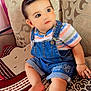 Erika joined the competition — help win amazing prizes! baby, child, toddler, denim_overalls, striped_shirt, couch, pillow, patterned, indoor, barefoot, cute, curly_hair, sitting, looking_away, infant, furniture, home, soft_lighting, portrait, skin