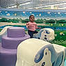 child, toddler, playground, soft_play, animal_shaped_seat, pink_sweater, indoor, net, life_ring, mural, colorful, wave, smile, fun, play_area, toy, floor, decor, casual_clothing, young_child