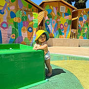 Nael a rejoint le concours — aidez-le/la à gagner de superbes lots ! child, toddler, baby, yellow_hat, splash_pad, water_fountain, colorful_mural, wooden_huts, outdoor, sunny, pool_area, swim_diaper, barefoot, standing, green_structure, shadow, playing, summer, curious, portrait