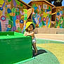 child, toddler, baby, yellow_hat, splash_pad, water_fountain, colorful_mural, wooden_huts, outdoor, sunny, pool_area, swim_diaper, barefoot, standing, green_structure, shadow, playing, summer, curious, portrait