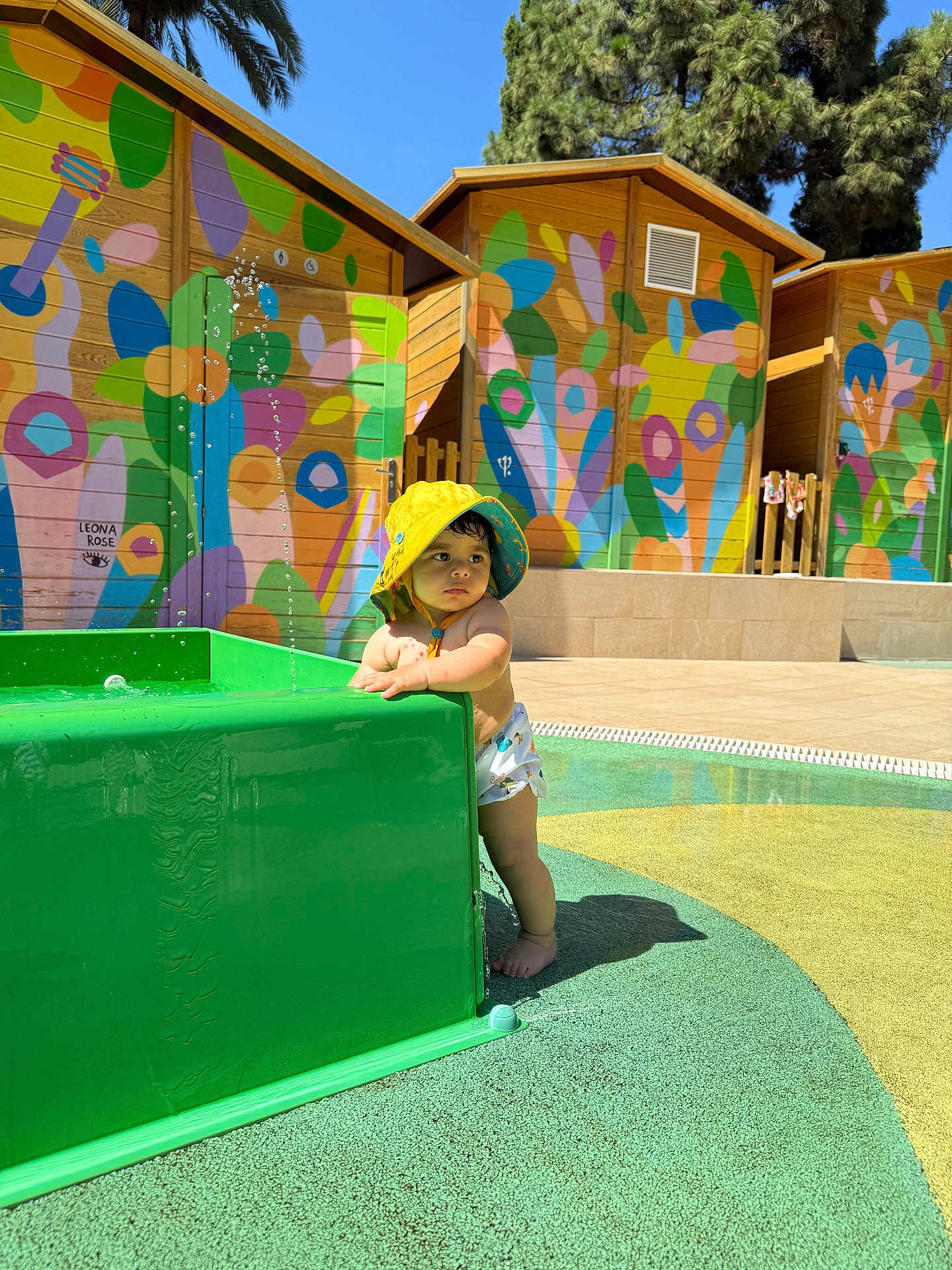 Nael a rejoint le concours — aidez-le/la à gagner de superbes lots ! child, toddler, baby, yellow_hat, splash_pad, water_fountain, colorful_mural, wooden_huts, outdoor, sunny, pool_area, swim_diaper, barefoot, standing, green_structure, shadow, playing, summer, curious, portrait