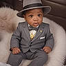 baby, child, suit, hat, fedora, bow_tie, jewelry, gold_necklace, couch, fur_cushion, portrait, indoors, cute, fashion, formal_wear, person, sitting, expression, big_eyes, sock