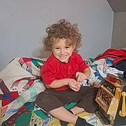 Luka is registered to the contest to win money with this photo: child, curly_hair, smiling, indoor, quilt, patchwork, red_shirt, dark_pants, black_socks, toy, slot_machine, playing, sitting, happy, bedroom, fabric, colorful, person, cute, cozy