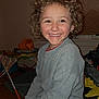 blanket, casual_clothing, child, cozy, curly_hair, curtain, fabric, floor, furniture, happy, home, indoor, long_sleeve_shirt, messy_face, person, portrait, sitting, smile, window, young