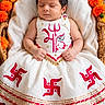 baby, infant, sleeping, portrait, wicker_basket, flowers, marigolds, traditional_clothing, white_dress, red_pattern, gold_trim, cultural_attire, indoors, peaceful, resting, close_up, blanket, decorative, photography, newborn
