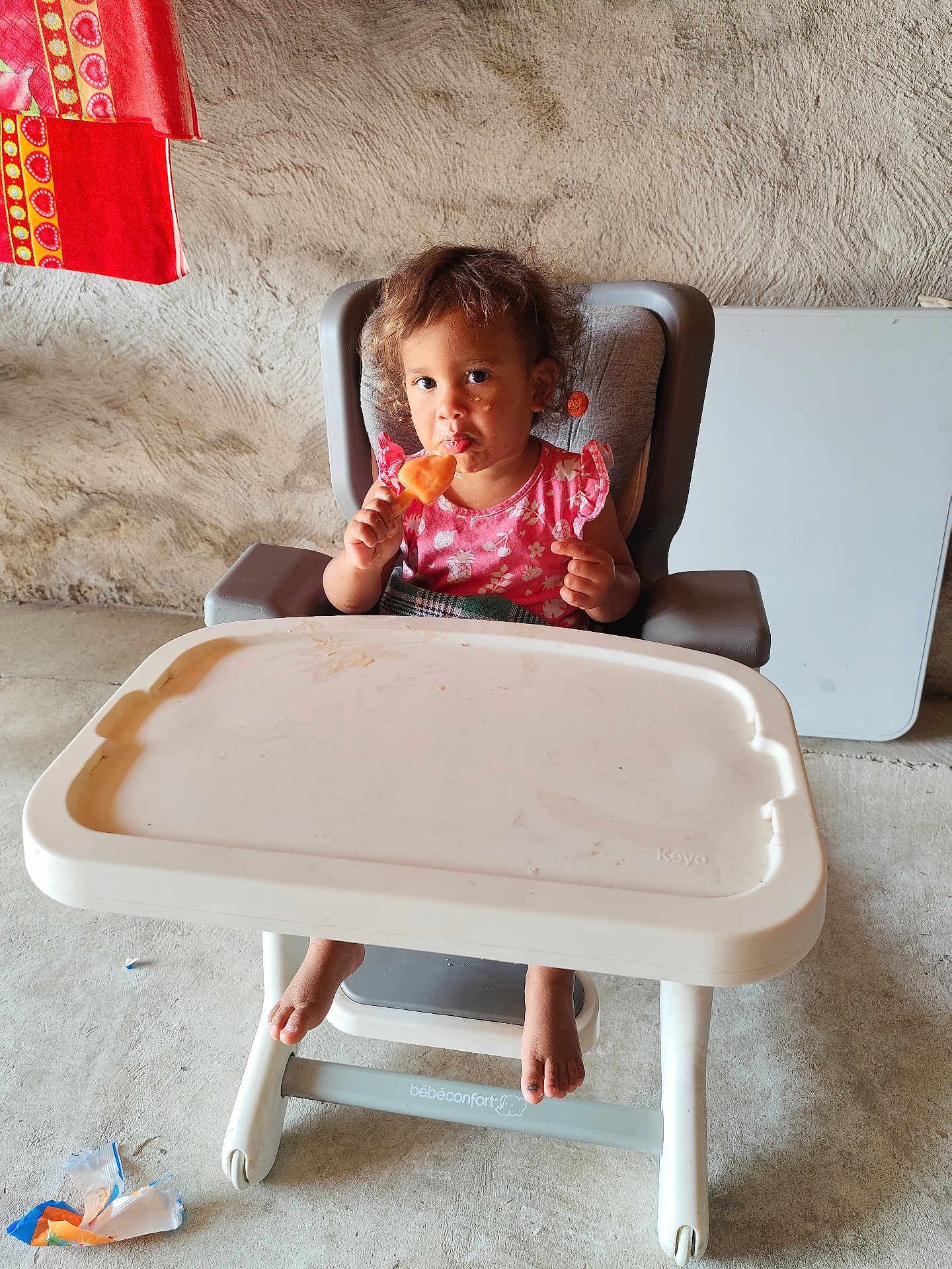 Maelle a rejoint le concours — aidez-le/la à gagner de superbes lots ! toddler, high_chair, popsicle, child, indoor, messy_face, pink_clothing, curly_hair, footwear_barefoot, plastic_tray, concrete_floor, textured_wall, colorful_cloth, toy_wrappers, cute, portrait, food, snack_time, little_feet, curious_expression