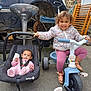 baby, car_seat, child, concrete, curly_hair, footwear, jacket, outdoor, pavement, person, pink_clothing, playtime, safety, seat_belt, smiling, toddler, toy, tricycle, vehicle, wheel