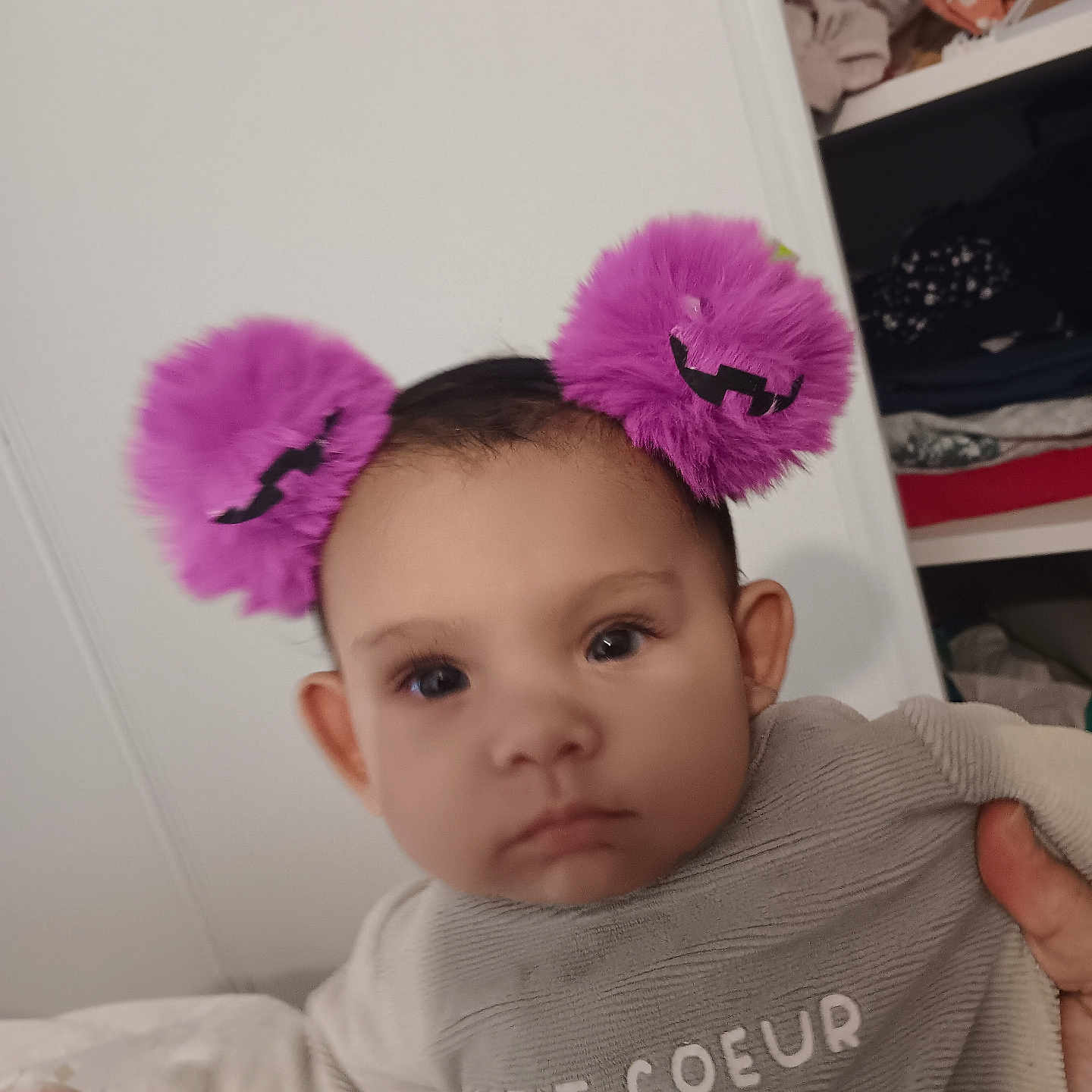 émiliana a rejoint le concours — aidez-le/la à gagner de superbes lots ! apparel, baby, child, closet, clothing, cute, expressionless, face, furniture, hand, headband, holding, indoor, infant, person, pom_pom, portrait, purple, sleeve, text_on_clothing