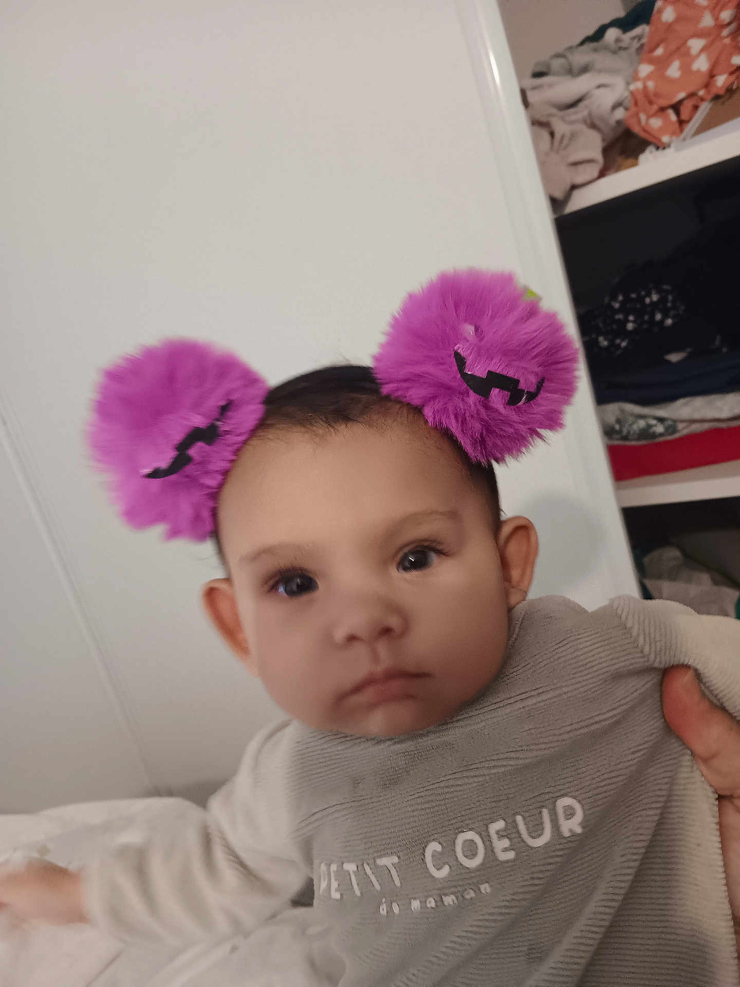 émiliana a rejoint le concours — aidez-le/la à gagner de superbes lots ! baby, child, pom_pom, headband, purple, clothing, face, person, indoor, closet, furniture, sleeve, hand, expressionless, infant, apparel, text_on_clothing, holding, portrait, cute