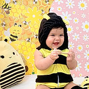 Alice is registered to the contest to win money with this photo: baby, child, costume, bee_costume, smiling, cute, happy, indoors, plush_toy, stuffed_animal, yellow, black, honeycomb, flowers, decor, background, sitting, playful, infant, headwear