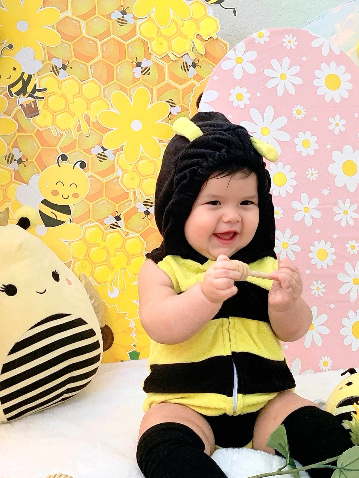Alice is registered to the contest to win money with this photo: baby, child, costume, bee_costume, smiling, cute, happy, indoors, plush_toy, stuffed_animal, yellow, black, honeycomb, flowers, decor, background, sitting, playful, infant, headwear