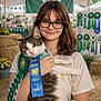 girl, cat, glasses, ribbon, award, four_h, county_fair, tent, bunting, rosette, hay_bales, straw, portrait, smile, pet, pride, booth, flowers, hands, background_people