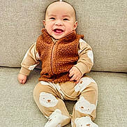 Andrew is registered to the contest to win money with this photo: baby, smiling, child, pajamas, bear_pattern, vest, brown_clothing, beige_couch, indoor, happy, cute, sitting, infant, face, portrait, cozy, soft_texture, clothing, person, young