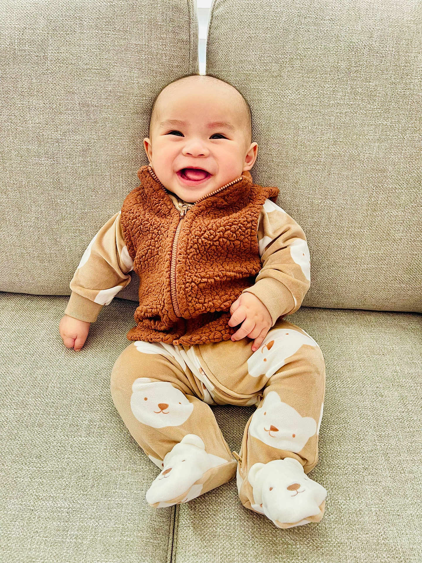 Andrew is registered to the contest to win money with this photo: baby, smiling, child, pajamas, bear_pattern, vest, brown_clothing, beige_couch, indoor, happy, cute, sitting, infant, face, portrait, cozy, soft_texture, clothing, person, young
