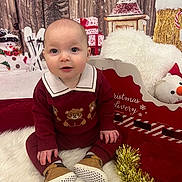Ellijah joined the competition — help win amazing prizes! baby, child, toddler, maroon_outfit, teddy_bear_pattern, brown_shoes, white_rug, christmas_decorations, snowman, wrapped_presents, wooden_backdrop, festive, holiday, tinsel_garland, plush_pillow, red_decor, smiling, sitting, indoor, cute
