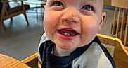 Ellijah is registered to the contest to win money with this photo: baby, child, high_chair, smiling, messy_face, indoor, table, wooden_furniture, shirt, long_sleeve, cafe, window, greenery, chair, face, person, teeth, happy, cute, infant