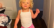 Ricki is registered to the contest to win money with this photo: child, toddler, curly_hair, blond, white_tank_top, pink_leggings, standing, barefoot, indoor, hallway, wooden_floor, door, laundry_basket, clothes, smiling, playful, person, young_child, cute, happy