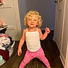 child, toddler, curly_hair, blond, white_tank_top, pink_leggings, standing, barefoot, indoor, hallway, wooden_floor, door, laundry_basket, clothes, smiling, playful, person, young_child, cute, happy