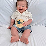 Leo is registered to the contest to win money with this photo: baby, child, smile, sitting, shirt, denim_shorts, wooden_sign, three_months, happy, indoors, couch, white, cute, portrait, infant, young_child, skin, feet, hand, face