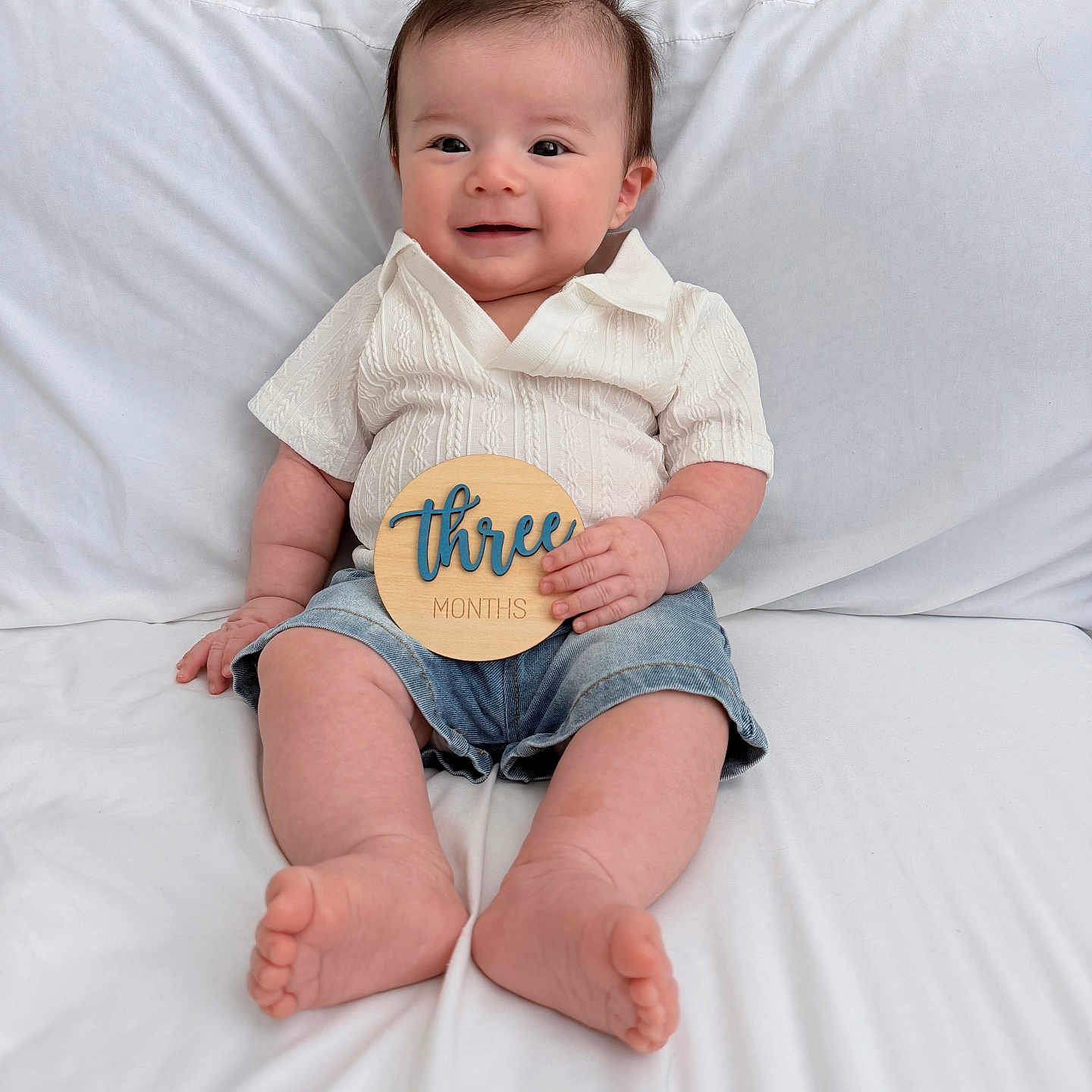 Leo is registered to the contest to win money with this photo: baby, child, smile, sitting, shirt, denim_shorts, wooden_sign, three_months, happy, indoors, couch, white, cute, portrait, infant, young_child, skin, feet, hand, face