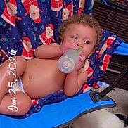 Josiah is registered to the contest to win money with this photo: baby, bottle, chair, blanket, santa_claus_pattern, dog, blue_collar, carpet, indoor, child, cute, reclining, drinking, furniture, person, curly_hair, skin, relaxing, pet, date_stamp