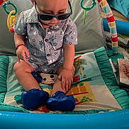 Aziel joined the competition — help win amazing prizes! baby, sunglasses, play_mat, toy, indoor, child, blue_shoes, shirt, star_pattern, floor, colorful, curious, seated, hand, head, face, clothing, footwear, baby_toys, playtime